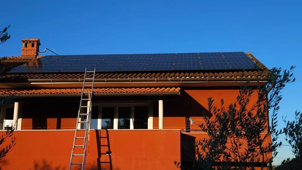 Široki pogled na solarne panele na crvenom popločanom krovu kuće, s plavim nebom i ljestvama uz pročelje.