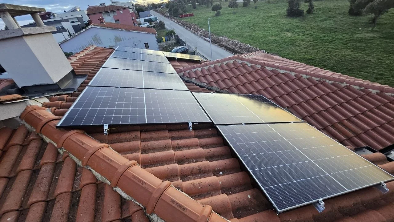 Solarni paneli na crvenom popločanom krovu uz dimnjak, s cestom i zelenom površinom u pozadini.